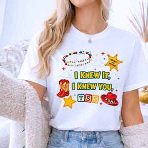 K&ouml;nnte beinhalten: Ein wei&szlig;es kurz&auml;rmeliges T-Shirt mit einem bunten Grafikdruck. Das Design zeigt ein Perlenarmband mit dem Text 'You've got a friend in me', einen gelben Sheriffstern, rote Cowboystiefel, einen roten Cowboyhut und Spielw&uuml;rfel mit der Aufschrift 'TS5'.