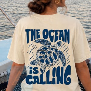 K&ouml;nnte beinhalten: Ein cremefarbenes Kurzarm-T-Shirt mit einer dunkelblauen Grafik auf der R&uuml;ckseite. Das Design zeigt eine Meeresschildkr&ouml;te, umgeben von wellenf&ouml;rmigen Linien, und den Text "THE OCEAN is CALLING" in einer fetten, stilisierten Schrift.