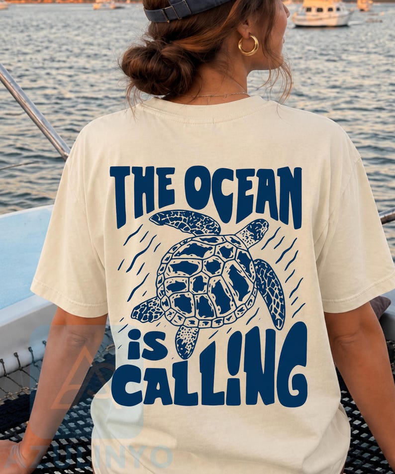K&ouml;nnte beinhalten: Ein cremefarbenes Kurzarm-T-Shirt mit einer dunkelblauen Grafik auf der R&uuml;ckseite. Das Design zeigt eine Meeresschildkr&ouml;te, umgeben von wellenf&ouml;rmigen Linien, und den Text "THE OCEAN is CALLING" in einer fetten, stilisierten Schrift.