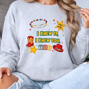 K&ouml;nnte beinhalten: Ein hellgraues Sweatshirt mit Rundhalsausschnitt und einem Grafikdruck mit dem Text I KNEW IT, I KNEW YOU, TS5 und INFINITY AND BEYOND. Das Design enth&auml;lt ein Perlenarmband, einen gelben Sheriffstern, rote Cowboystiefel, einen roten Hut und einen gelben Stern.