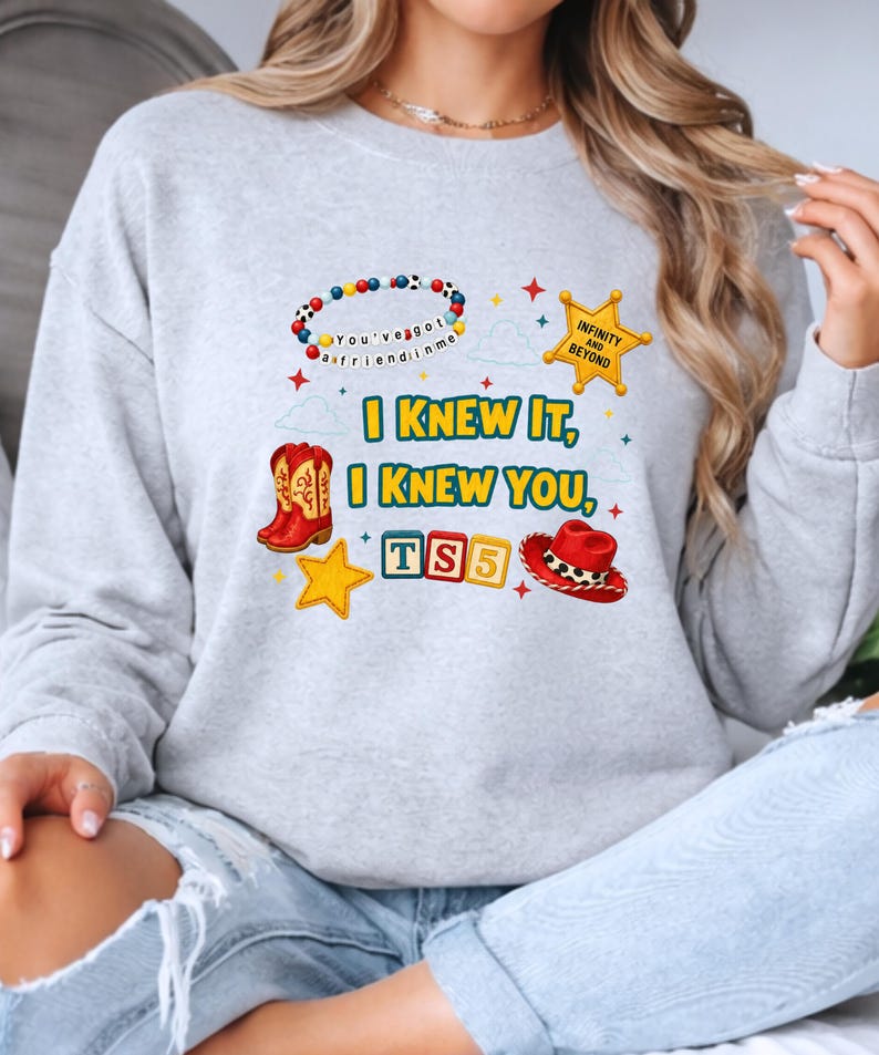 K&ouml;nnte beinhalten: Ein hellgraues Sweatshirt mit Rundhalsausschnitt und einem Grafikdruck mit dem Text I KNEW IT, I KNEW YOU, TS5 und INFINITY AND BEYOND. Das Design enth&auml;lt ein Perlenarmband, einen gelben Sheriffstern, rote Cowboystiefel, einen roten Hut und einen gelben Stern.
