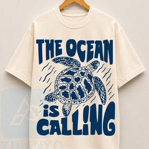 K&ouml;nnte beinhalten: Ein cremefarbenes Kurzarm-T-Shirt mit einer dunkelblauen Grafik einer schwimmenden Meeresschildkr&ouml;te. Der Text "THE OCEAN is CALLING" ist in einer fetten, dunkelblauen Schrift &uuml;ber und unter der Schildkr&ouml;tenillustration abgebildet.