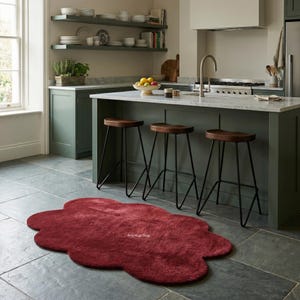 May include: A burgundy cloud-shaped rug on a slate tile floor in a kitchen. The rug is in front of a kitchen island with three wooden stools. The kitchen has green cabinets and shelves with dishes. The rug is a decorative floor covering.