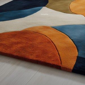 May include: Close-up of a colorful area rug with an abstract design. The rug features large, overlapping shapes in shades of orange, blue, beige, and green. The rug is made of a soft, textured material and is placed on a light-colored wooden floor.