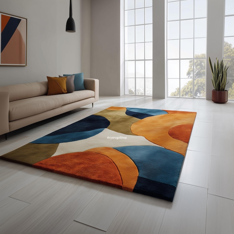 May include: A rectangular area rug with an abstract design in shades of orange, blue, brown, and cream. The rug features a modern, geometric pattern with overlapping shapes. The rug is placed on a light-colored floor in a living room setting.