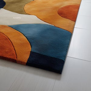 May include: A colorful area rug with an abstract design. The rug features a combination of orange, blue, white, and green shapes. The rug's texture is visible, and the design is modern and eye-catching.