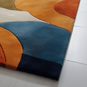 May include: Close-up of a colorful area rug with an abstract design. The rug features a combination of orange, blue, cream, and brown hues. The rug's texture is visible, and the design is reminiscent of a landscape.