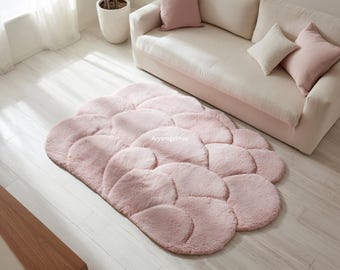 May include: A soft, pink cloud-shaped rug with a plush texture. The rug is placed on a light wood floor in front of a beige sofa with pink pillows. The rug's design features overlapping, rounded shapes.