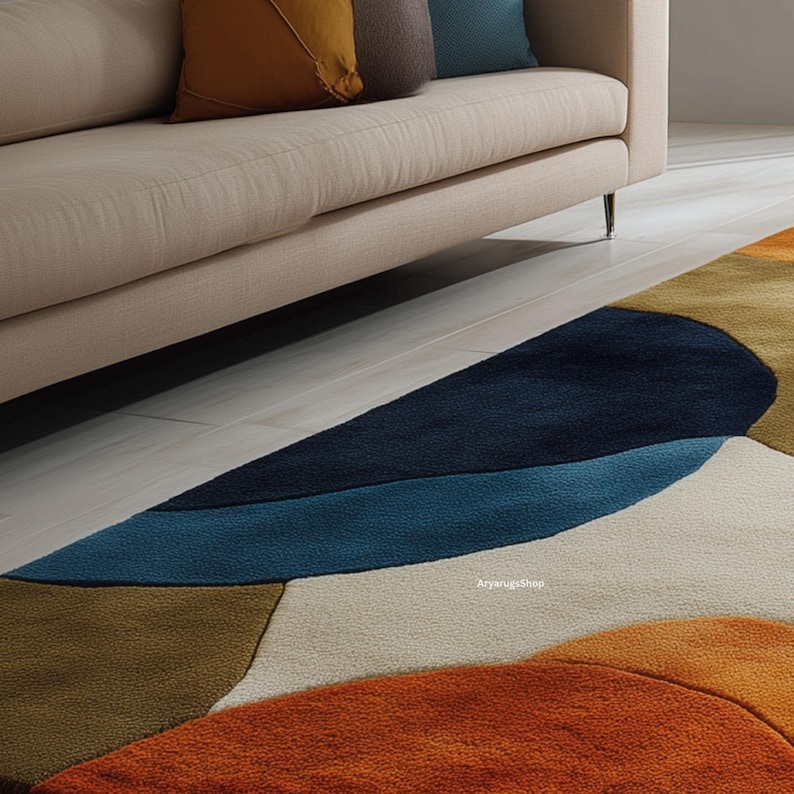 May include: A modern area rug with an abstract design. The rug features a combination of colors, including orange, brown, blue, and cream. The rug is placed on a light-colored wooden floor, with a beige sofa and pillows in the background.