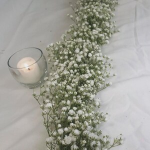 May include: A table centerpiece featuring a long garland of white baby's breath flowers. Two glass votive candle holders with lit candles are placed on either side of the floral arrangement, creating a soft, romantic ambiance.