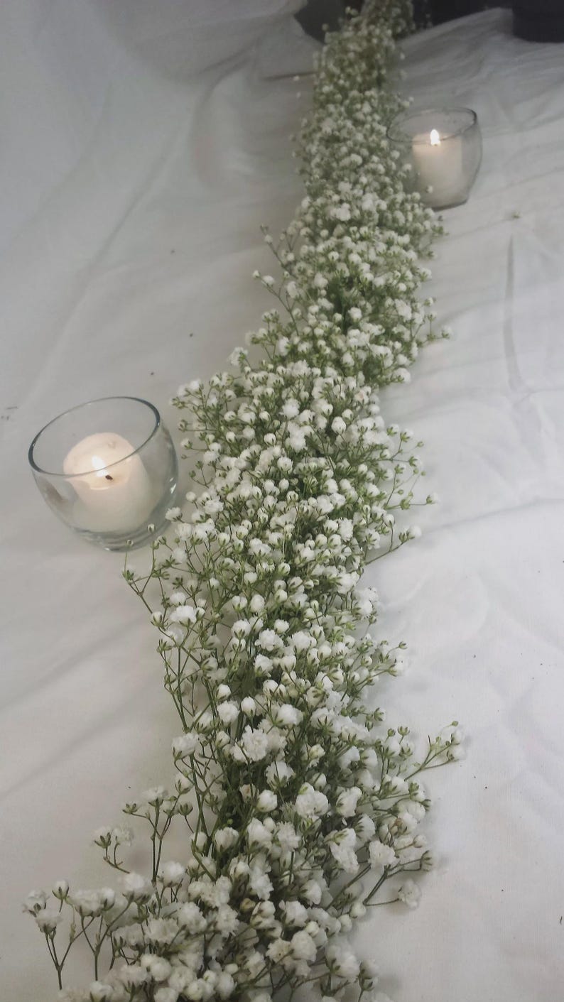 May include: A table centerpiece featuring a long garland of white baby's breath flowers. Two glass votive candle holders with lit candles are placed on either side of the floral arrangement, creating a soft, romantic ambiance.