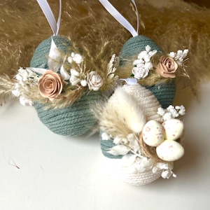 Op de afbeelding: Drie decoratieve paasei-ornamenten. Twee zijn blauwgroen en één is wit, allemaal met witte linten om op te hangen. Elk ei is versierd met gedroogde bloemen, kunstrozen en kleine gespikkelde eieren. De eieren zijn ontworpen voor Pasen.