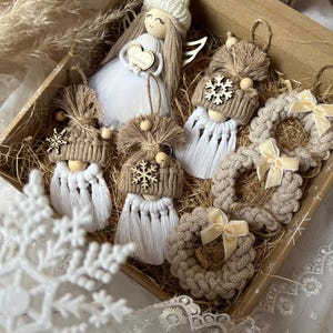 Op de afbeelding: Een verzameling handgemaakte kerstversieringen in een kartonnen doos. De ornamenten omvatten een witte engel met een houten hart, kabouterachtige figuren met gebreide mutsen en gevlochten kransen met crèmekleurige strikken. Het kleurenpalet is neutraal met beige, wit en bruin.