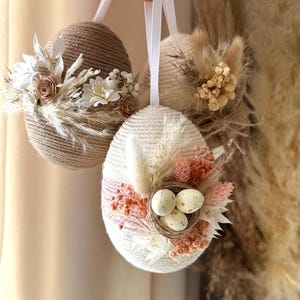 Può includere: Tre decorazioni a forma di uovo di Pasqua. Le uova sono avvolte in spago e filo, decorate con fiori secchi, piume e piccoli nidi con uova macchiate. Ogni uovo ha un nastro bianco per appenderlo. La tavolozza dei colori comprende beige, bianco e rosa.