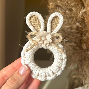 Puede incluir: Adorno en forma de conejito tejido de color crema, con orejas de arpillera y un adorno floral. El conejito tiene un cuerpo circular y está decorado con hilo. Decoración temática de Pascua.