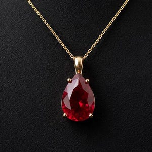 Könnte beinhalten: Eine goldene Halskette mit einem großen, facettierten, birnenförmigen roten Edelsteinanhänger. Der leuchtend rote Stein ist in einer einfachen goldenen Fassung mit vier Krappen befestigt und hängt an einer zarten goldenen Gliederkette.