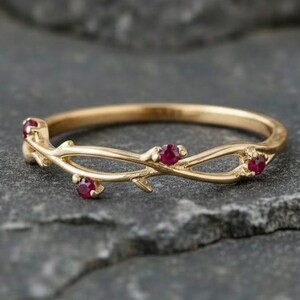 May include: A gold ring featuring a band designed to resemble a thin, intertwining twig or branch. The ring is set with three small, round, vibrant red rubies placed at intervals along the textured, organic-looking metal surface of the band.