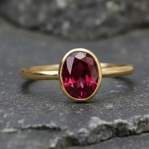 Könnte beinhalten: Ein goldener Ring mit einem großen, oval geschliffenen, tiefroten Edelstein. Der Edelstein ist in einer Zargenfassung gefasst, und das Band ist schlichtes, poliertes Gold. Der Ring wird auf einer dunkelgrauen, strukturierten Oberfläche gezeigt.
