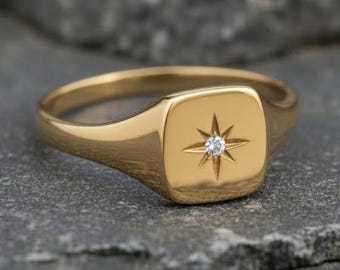 Puede incluir: Un anillo de sello dorado con una cara cuadrada. La cara presenta un diseño de estrella con una pequeña gema redonda y transparente en el centro. El anillo está sobre un fondo gris texturizado.
