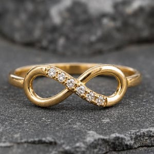 May include: A gold ring featuring an infinity symbol design. One side of the infinity loop is embellished with five small, clear, round-cut stones set into the metal band. The ring has a polished finish and a continuous, smooth, circular band structure.