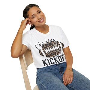 May include: White t-shirt with a football graphic featuring a leopard print design. The shirt has the words "Classy" and "Kickoff" printed on it in black script. The model is wearing blue jeans and sitting on a wooden chair.