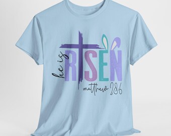Könnte beinhalten: Hellblaues T-Shirt mit einem violetten Kreuz und den Worten "He is Risen" in bunten Buchstaben. Der Text "Matthew 28:6" steht unter dem Kreuz.