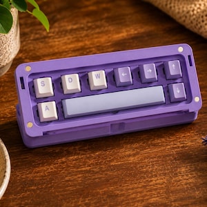 Peut inclure: Un pavé macro pour clavier mécanique rectangulaire violet avec une barre d'espace violet clair et des touches blanches marquées de lettres violettes, dont S, D, W et des flèches directionnelles. Le boîtier violet comporte quatre accents métalliques.