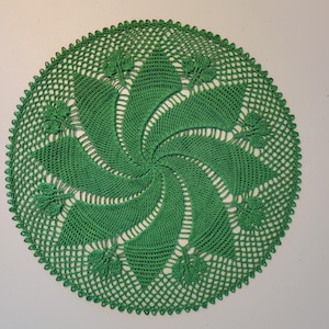 May include: A handmade, circular green doily with a spiral design. The center features a tightly crocheted spiral, surrounded by openwork and decorative leaf-like accents along the edge. The doily is approximately 12 inches in diameter.