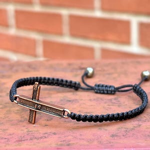 May include: A black braided cord bracelet featuring a central copper-toned metal cross charm. The bracelet includes an adjustable sliding knot closure with two small metallic beads at the ends of the cords, providing a secure and flexible fit.