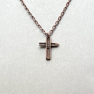 May include: A copper-toned metal cross pendant suspended from a matching link chain. The cross features a textured, weathered surface with a vertical bar and a shorter horizontal bar, creating a simple, industrial-inspired design with a dark finish.