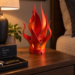 May include: A sculptural lamp shaped like a stylized flame with a vibrant red and orange gradient finish. The object features multiple upward-curving, pointed flame-like extensions rising from a circular base, creating a dynamic, layered appearance.