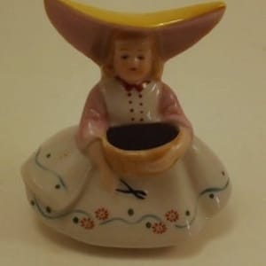 May include: A porcelain figurine of a girl wearing a yellow hat and a pink dress, holding a basket with a pair of scissors. The figurine is a vintage trinket or decorative piece.