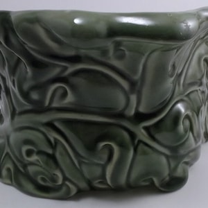 May include: A green ceramic planter with an intricate, swirling design. The planter is shaped like a bowl with a wide opening and a rounded bottom.