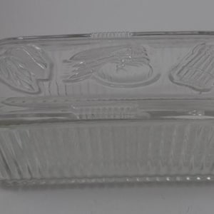 May include: A clear glass rectangular dish with a lid. The lid has a raised design of vegetables, including an onion, a tomato, and asparagus.