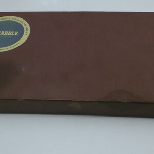 May include: A brown cardboard box with a circular label that says "SCRABBLE" in gold letters. The box is closed and appears to be a game box.