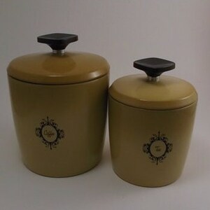 May include: Two vintage ceramic canisters with black lids. The larger canister is labeled "Coffee" and the smaller canister is labeled "Tea". Both canisters are a muted yellow color.