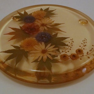 May include: A round, clear, glass trivet with a pressed flower design. The flowers are in shades of purple, yellow, and white. The trivet has a brown rim.