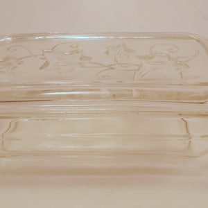 May include: A clear glass butter dish with a lid. The lid has a raised design of three stylized figures.