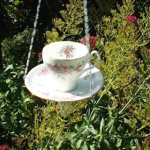 May include: A white teacup and saucer with a floral pattern hanging from a beaded chain. The teacup and saucer are suspended in the air, with the teacup facing the camera.
