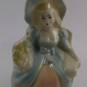 May include: A ceramic figurine of a woman wearing a blue and white dress and a hat. She is holding a bouquet of flowers in her hands.