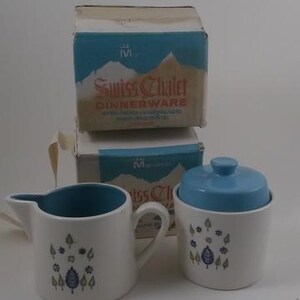 May include: A set of white ceramic creamer and sugar bowl with blue lids. The bowls have a blue and green floral pattern with a white background. The boxes are labeled "Swiss Chalet Dinnerware".