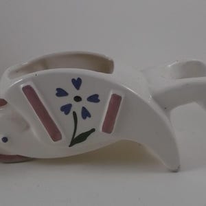 May include: White ceramic planter shaped like a wheelbarrow with a pink and purple flower design.