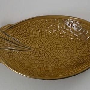 May include: A large, oval-shaped ceramic serving dish with a textured, mosaic-like design in shades of brown. The dish has a unique, leaf-shaped handle on one side.