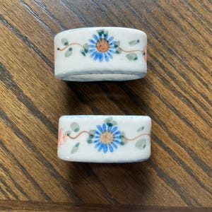 May include: Two white ceramic napkin rings featuring a hand-painted floral pattern. Each ring displays a central blue daisy-like flower with a tan center, surrounded by thin brown stems, small green leaves, and soft pink accents on a glossy surface.