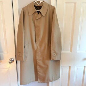 May include: A tan trench coat featuring a wide collar, long sleeves with buttoned cuffs, and a concealed front button placket. The coat has a smooth, solid-colored fabric construction with a dark brown interior lining visible at the neckline.