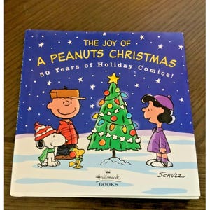 May include: A Peanuts Christmas book cover with a blue background and white stars. The cover features Charlie Brown, Snoopy, Lucy, and a Christmas tree. Text reads "The Joy of A Peanuts Christmas" and "50 Years of Holiday Comics!"