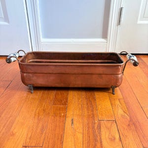 May include: A rectangular copper planter with a smooth finish, featuring two side handles with white and blue floral ceramic accents. The vessel is supported by four small metal feet and has an open top with a deep interior basin.