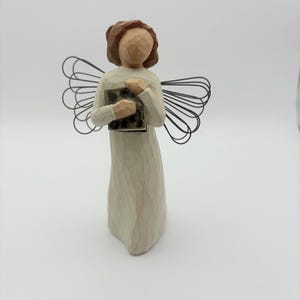 May include: A standing figurine of an angel with brown hair, wearing a long, off-white textured gown. The figure holds a small, dark, rectangular object in its hands and features large, decorative wings made of thin, black, looped wire.