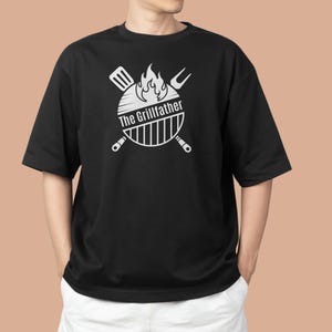 Peut inclure: T-shirt noir avec un motif blanc représentant un barbecue, des flammes, une spatule et une fourchette. Le texte "The Grillfather" est centré dans le motif. T-shirt décontracté à manches courtes.