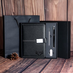 May include: A black leather journal with a silver clasp, a pen, and a pen refill are displayed in a black gift box. The journal has the initials "GH" and a deer antler design. A black gift bag and a pine cone are also present.